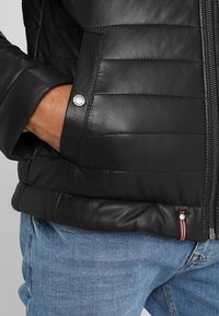 Hand i fickan på en svart vadderad läderjacka, synlig tryckknapp, dragkedja och randig tygetikett; person med blå jeans.