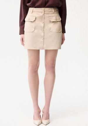 Beżowa mini spódnica z przodu zapinana na guziki, z dwoma dużymi klapkowymi kieszeniami z przodu, noszona z brązową bluzką z długim rękawem i beżowymi szpilkami.