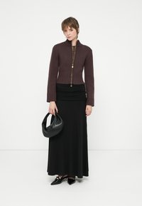 Žena u smeđem zip-up dukserici, crnoj maksi suknji s naborima, crnim cipelama s ukazom, drži crnu kožnu torbu na bijeloj pozadini.
