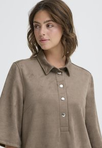 Ruda veliūrinė marškiniai su apykakle, trumpomis rankovėmis ir sidabriniais spaudžiamais mygtukais per visą priekinę dalį. Minkšta tekstūra, minimalus dizainas.