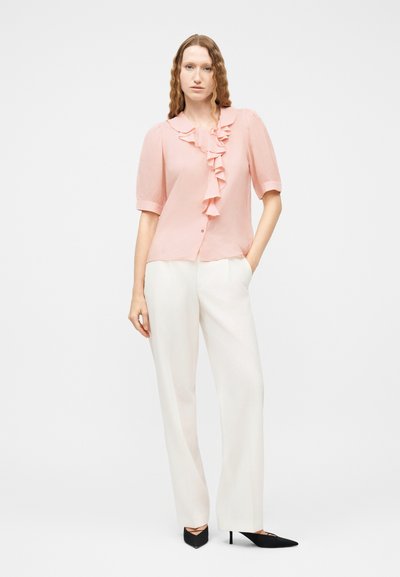 Femme portant un chemisier rose à manches courtes avec des volants et un pantalon blanc à jambes larges, debout avec une main dans la poche et des talons noirs.
