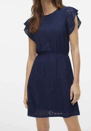 Marineblauwe eyelet-jurk met opstaande mouwen, getailleerde taille en diamantvormige uitsnijdingen, gedragen door een staande vrouw.