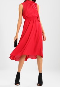 Rode mouwloze jurk van vloeiende stof, met een getailleerde taille en een asymmetrische zoom, gecombineerd met zwarte open-toe enkellaarsjes en een clutch.