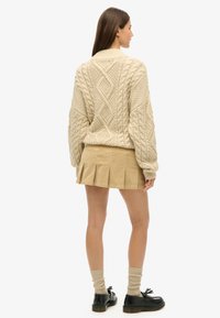 Beige cable-strikket sweater med høj krave, parret med en tan plisseret nederdel. Båret med knæhøje ribbede strømper og sorte snørebåndssko.