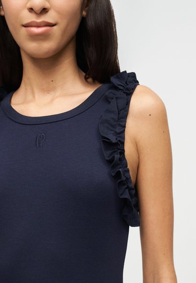 Γυναίκα που φοράει μπλε σκούρο αμάνικο τοπ με λεπτομέρεια από βολάν στο ύψος του ώμου και ένα μικρό κεντημένο λογότυπο στο στήθος.