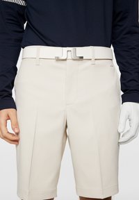 Beżowe szorty z bocznymi kieszeniami, gładka tekstura i pasek z metalową klamrą w kolorze srebrnym. W połączeniu z ciemną koszulą z długim rękawem i białą rękawiczką.