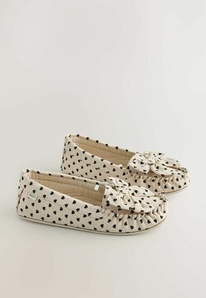 Beige fabric slippers with black heart pattern, bow accent on the front, soft texture, rounded shape, and gathered stitching detail.
