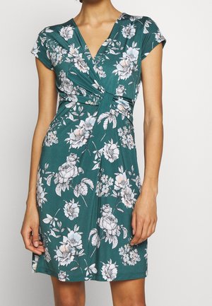 Femme portant une robe verte mi-longue à manches courtes avec un motif floral blanc et rose clair et un détail froncé torsadé à la taille.