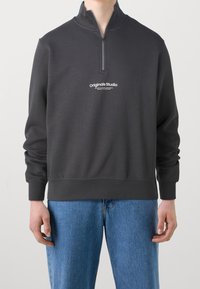 Uomo che indossa una felpa nera con zip a quarto e logo "Originals Studio" e jeans blu, in piedi contro uno sfondo bianco neutro.