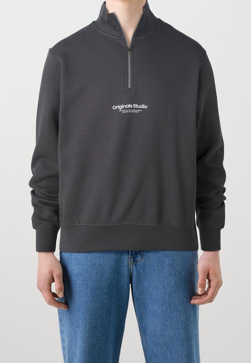 Uomo che indossa una felpa nera con zip a quarto e logo "Originals Studio" e jeans blu, in piedi contro uno sfondo bianco neutro.
