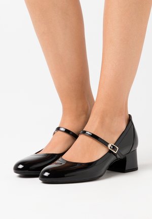 Musta patentoitu nahka Mary Jane -kengät matalilla paksuilla kengänpohjilla, käytettyinä paljain jaloin, esitettynä valkoista taustaa vasten.