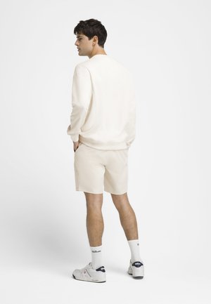 Mann trägt cremefarbenen Sweatshirt und Shorts mit weißen Wilson-Socken und Schuhen, steht seitlich mit Händen in den Taschen.