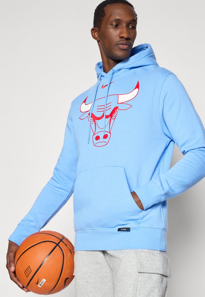 Helle blaue Kapuzenjacke aus Baumwollmischung, mit einem roten Bullenmotiv auf der Brust und einer Kängurutasche. Modell hält einen Basketball.