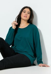 Een teal gekleurde langwerpige mouwshirt van zachte stof met een ronde hals, gecombineerd met zwarte slim-fit jeans, zittend voor een witte achtergrond.