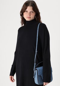 Borsa a tracolla in suede blu con tracolla regolabile, forma quadrata, texture morbida e chiusura a patta. Il modello indossa un abito a collo alto nero.