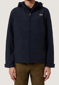 Man i en marinblå huva jacka med framtida dragkedja och sidofickor, kombinerad med bruna byxor, mot en enhetlig bakgrund.