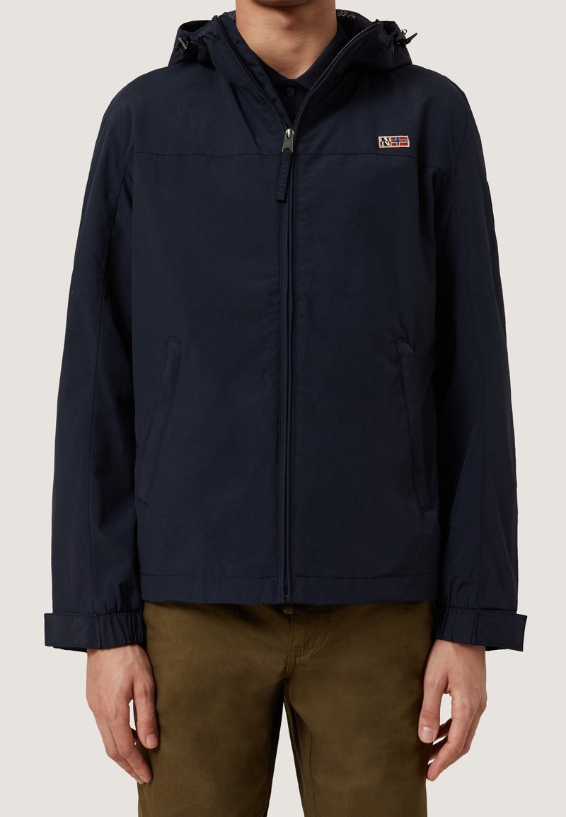 Man i en marinblå huva jacka med framtida dragkedja och sidofickor, kombinerad med bruna byxor, mot en enhetlig bakgrund.