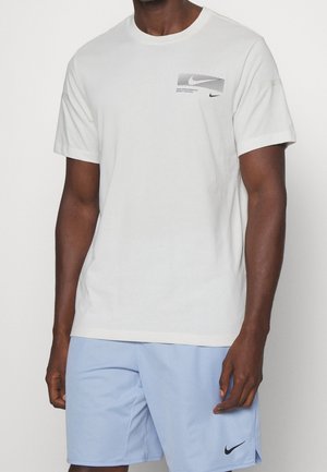 Mężczyzna ubrany w białą koszulkę Nike z logo i jasnoniebieskie szorty Nike, stoi na tle gładkiego białego tła.