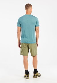 Heren t-shirt in een relaxte pasvorm in teal, lichte groene shorts met een achterzak; gecombineerd met zwart-geel wandelschoenen, op een effen achtergrond.
