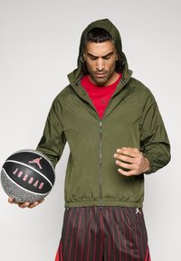 Λαδί πράσινο μπουφάν με κουκούλα και φερμουάρ, υφασμένο με ύφασμα με υφή, φορεμένο πάνω από κόκκινο πουκάμισο· κρατάει μια μαύρη και  γκρι μπάλα του μπάσκετ με κόκκινες λεπτομέρειες.