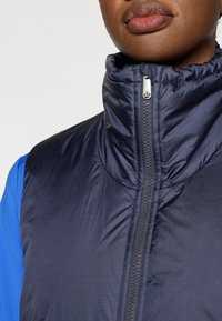 Colete acolchoado azul-marinho com gola alta, apresentando fecho de correr frontal e acabamento texturizado. Material leve com brilho suave.