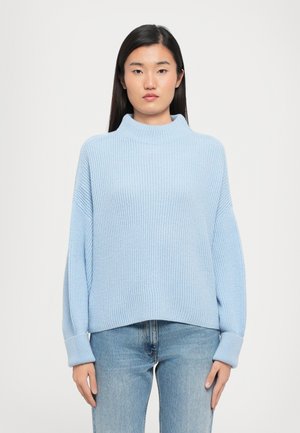 Lyseblå ribbet genser med høy hals og vide ermer, vist over tettsittende blå jeans. Teksturen virker myk og strikket.