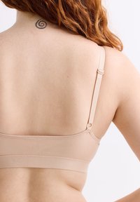 Roodharig persoon met een beige bh, die de achterkant laat zien met een verstelbare band en een spiraaltattoo op de bovenkant van de nek.