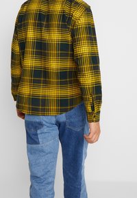 Geel en donkergroen geruit overhemd met lange mouwen, gestyled met blauwe spijkerbroek met gemengde texturen en kleuren. Knopenmanchetten zichtbaar.