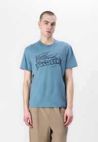 Niebieska bawełniana koszulka z okrągłym dekoltem i krótkimi rękawami, z ciemnoniebieskim logo Lacoste w kształcie krokodyla. Noszona z beżowymi spodniami.