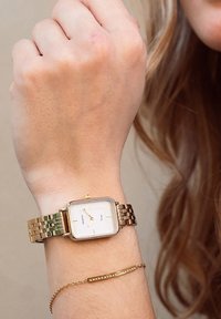 Montre carrée dorée avec un cadran blanc, trois aiguilles dorées et un bracelet à maillons. Dotée d'un délicat bracelet en chaîne dorée.