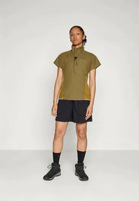 Kvinna står klädd i en olivgrön halvdraget kortärmad tröja, svarta shorts med fickor, svarta strumpor och svarta vandringskängor på grått golv.