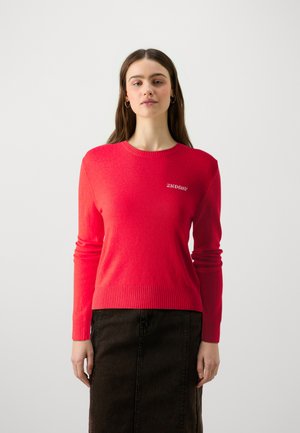 Red knitted sweater with ribbed hem and cuffs, featuring a small white "2ND DAY" logo on the chest. Paired with a dark skirt.