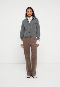 Kobieta stoi w szarym kardiganie zapinanym na guziki, nosząc paskowaną koszulę, brązowe spodnie z korduroy i białe buty, na tle gładkiego białego tła.