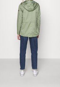 Persona con una giacca con cappuccio verde chiaro, jeans blu scuri e sneaker bianche alte, in piedi di fronte a una parete bianca uniforme.
