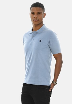 Junger Mann trägt ein hellblaues Poloshirt mit dunklem Logo, schwarze Hose und eine Armbanduhr, stehend vor einem schlichten weißen Hintergrund.