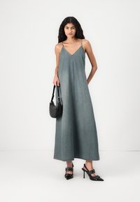 Denim maxi-kjole i falmet blå med V-utringning og tynne stropper, parret med svarte spisse hæler og en teksturert svart håndveske.
