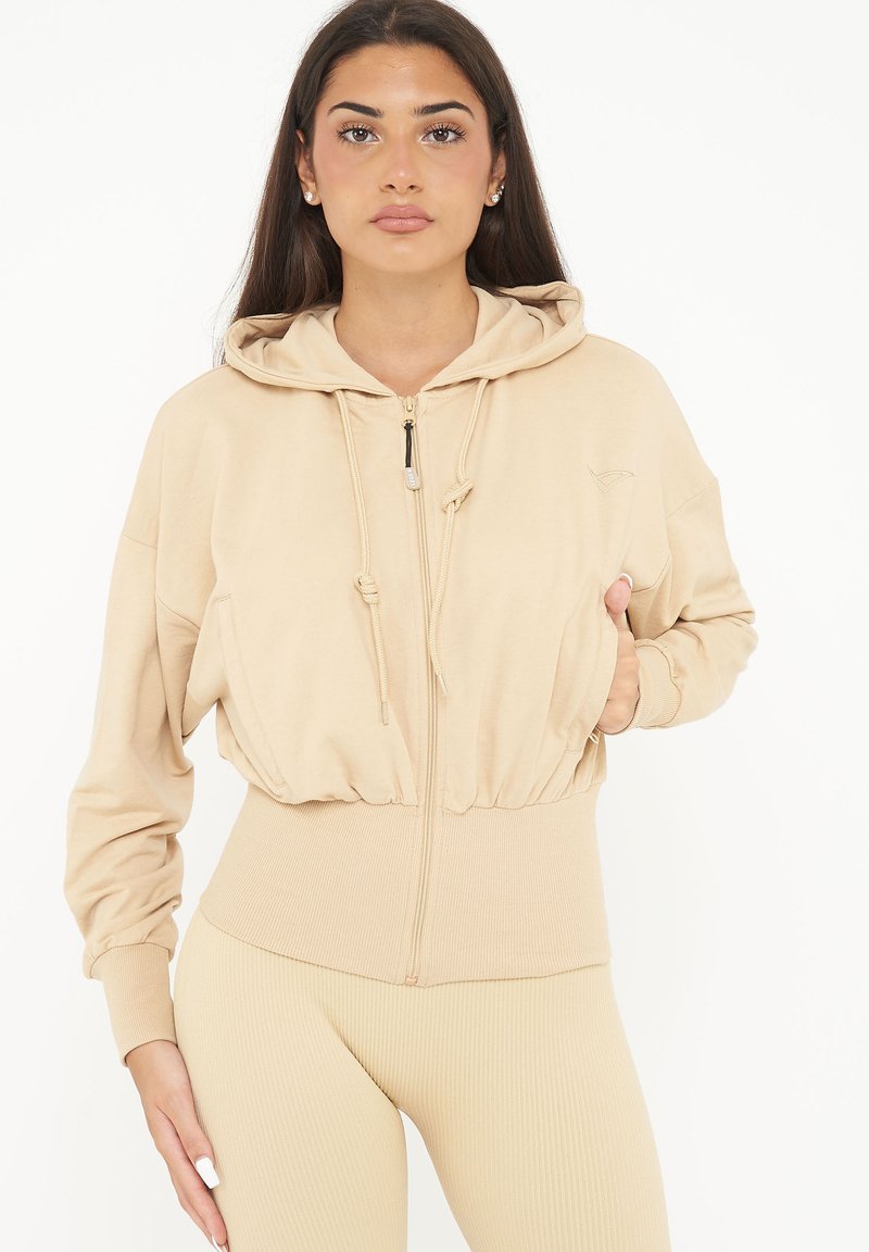 Sudadera beige con cremallera, capucha ajustable, dobladillo acanalado y bolsillos laterales; tejido suave con un corte relajado. Llevada con leggings a juego.