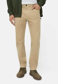 Beige Baumwollhose mit geradem Bein, detailreicher Nahtführung, zwei Eingrifftaschen und Knopfverschluss. Kombiniert mit dunkelgrünen Schuhen.