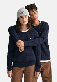 Marineblaue, kabelgestrickte Pullover mit gerippten Bündchen und Saum, über weißen Hemden getragen. Eine graue Mütze mit einem Logo. Der Hintergrund ist neutral und schlicht.