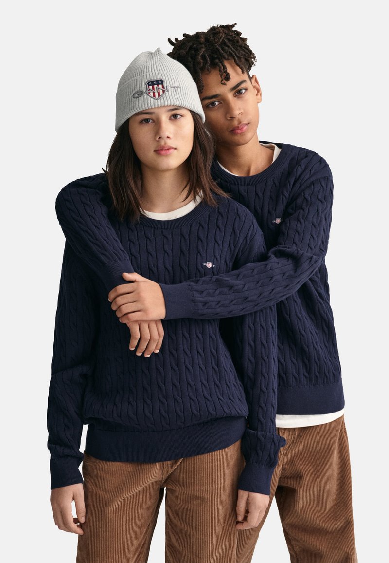 Marineblaue, kabelgestrickte Pullover mit gerippten Bündchen und Saum, über weißen Hemden getragen. Eine graue Mütze mit einem Logo. Der Hintergrund ist neutral und schlicht.
