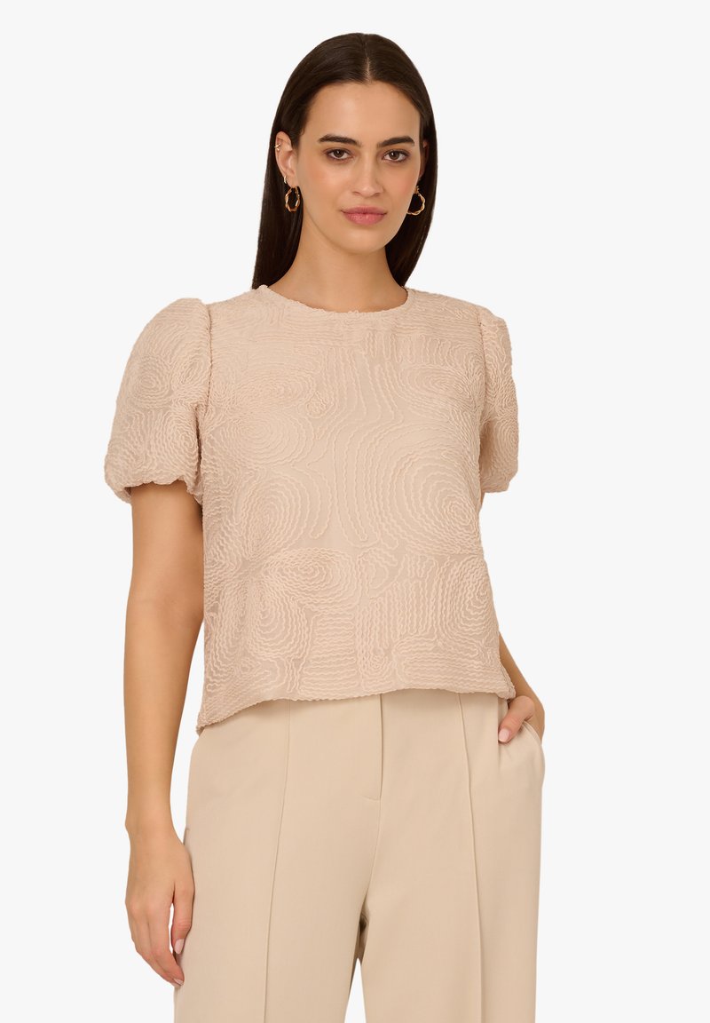 Haut en beige clair à texture avec des manches bouffantes, motif en vagues, encolure ronde et coupe décontractée, associé à un pantalon crème.
