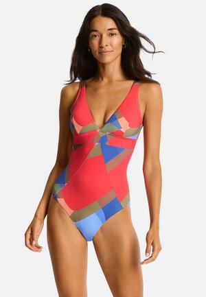 Donna che indossa un costume intero colorato con motivo geometrico con forme rosse, blu, marroni e rosa, in piedi su sfondo bianco.