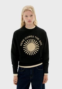 Čierny crewneck sveter s bledohnedou grafikou slnka a textom "HERE COMES THE SOL". Má rebrované manžety a lem. Voľný strih.