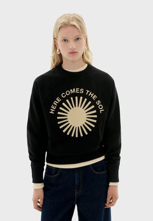 Felpa nera a girocollo con una grafica solare beige chiaro e il testo "QUI VIENE IL SOLE". Presenta polsini e orlo a costine. Vestibilità comoda.