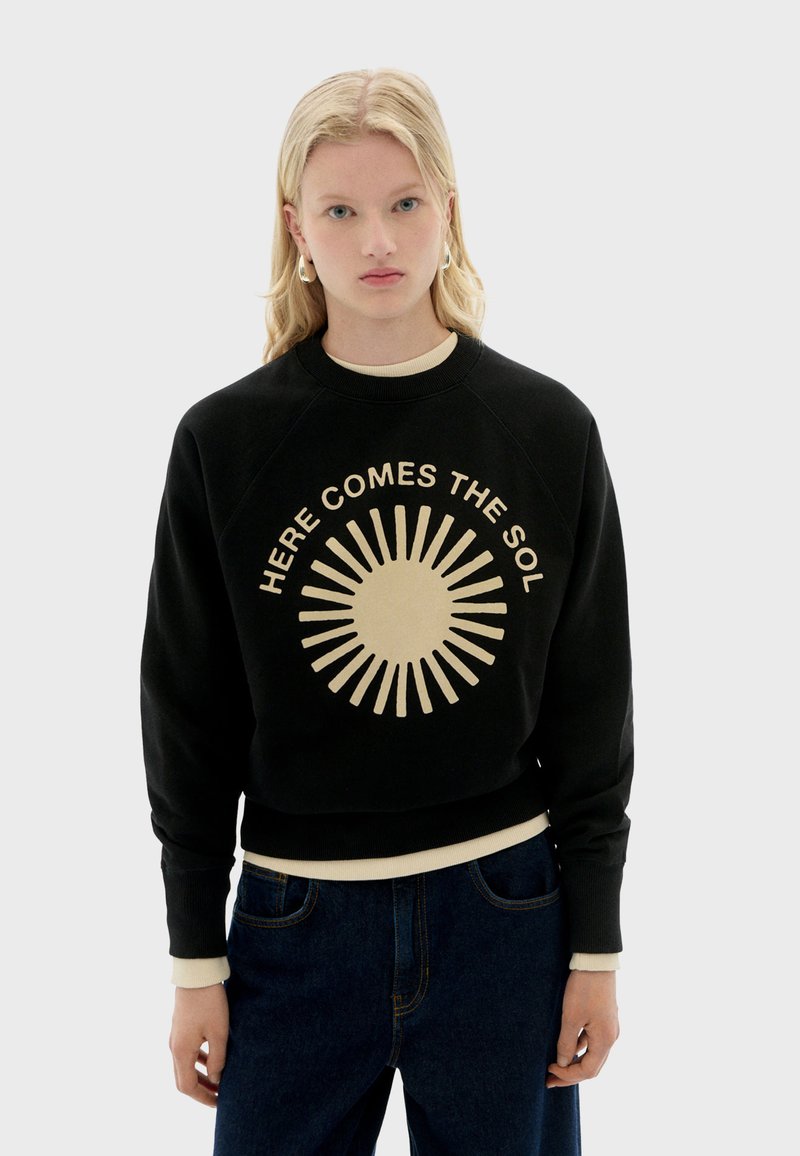 Sweatshirt noir à col ras du cou avec un graphique de soleil beige clair et le texte "Ici vient le soleil". Il présente des poignets et un ourlet côtelés. Coupe décontractée.