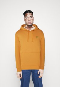 Haut à capuche orange en tissu doux, doté d'une poche kangourou et d'un logo brodé sur la poitrine. Associé à un pantalon bleu.