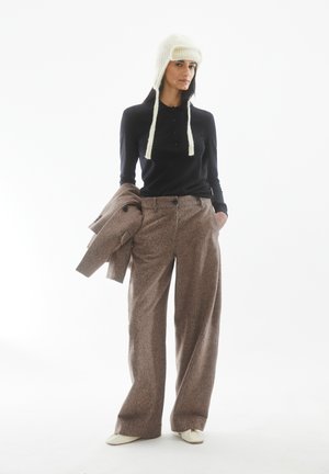 Braune Woll-Hose mit weitem Bein, schwarzes Langarmtop, cremefarbene Strickmütze mit Bändern, hält einen passenden braunen Mantel. Schlichtes, funktionales Design.