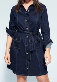 Robe chemise en denim foncé avec des manches longues, boutons sur le devant, ceinture à la taille et deux poches avant. Présente des coutures contrastées et des poignets de manches roulés.