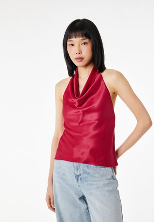 Femme aux cheveux noirs et raides portant un haut dos nu en satin rouge et un jean taille haute bleu clair sur fond blanc.
