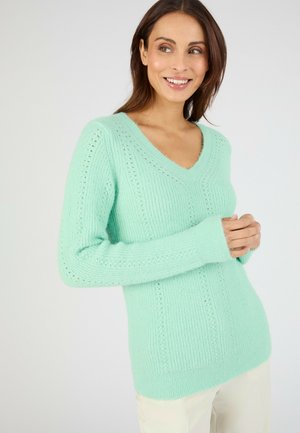 Frau mit dunklen Haaren, die einen mintgrünen, gestrickten V-Ausschnittpullover und hellfarbene Hosen trägt, lächelt und schaut leicht zur Seite.